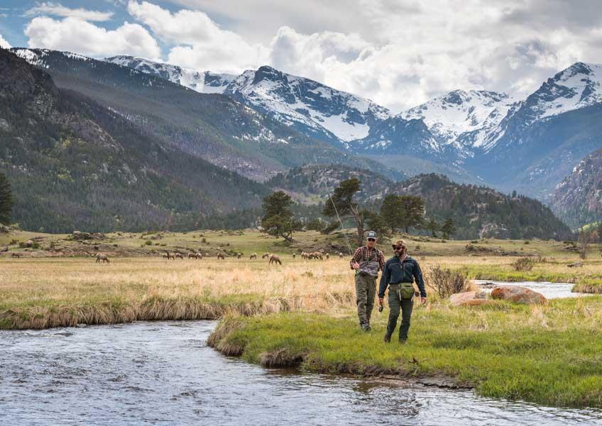 Two fisherman walk along a green-grass covered riverbank next to a cascading river. On the left bank, yellow tall grasses sway with elk grazing. In the distance snow-capped mountain peaks meet a cloudy sky.