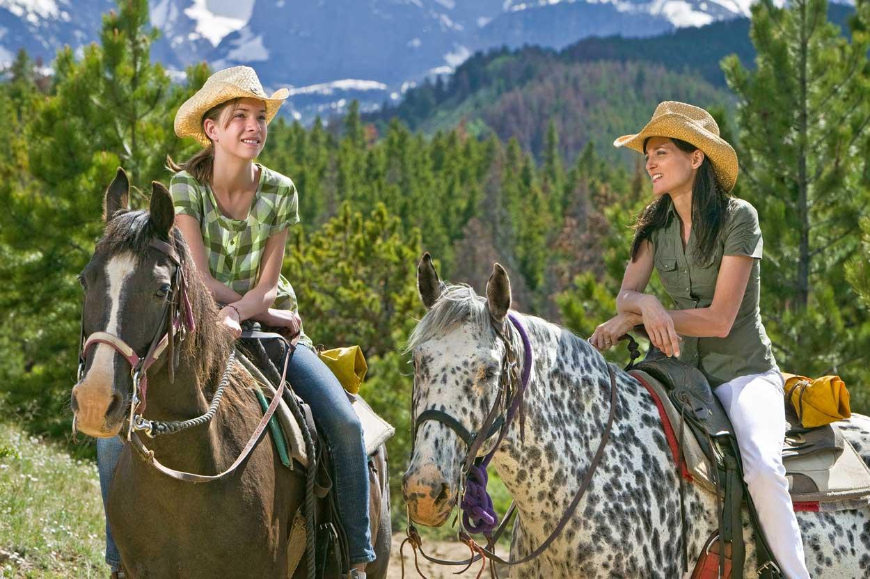 Two women sit atop horses — one horse is brown and the other is white with black spots — on a summer's day with evergreen trees on hills with a mountain peak in the distance.