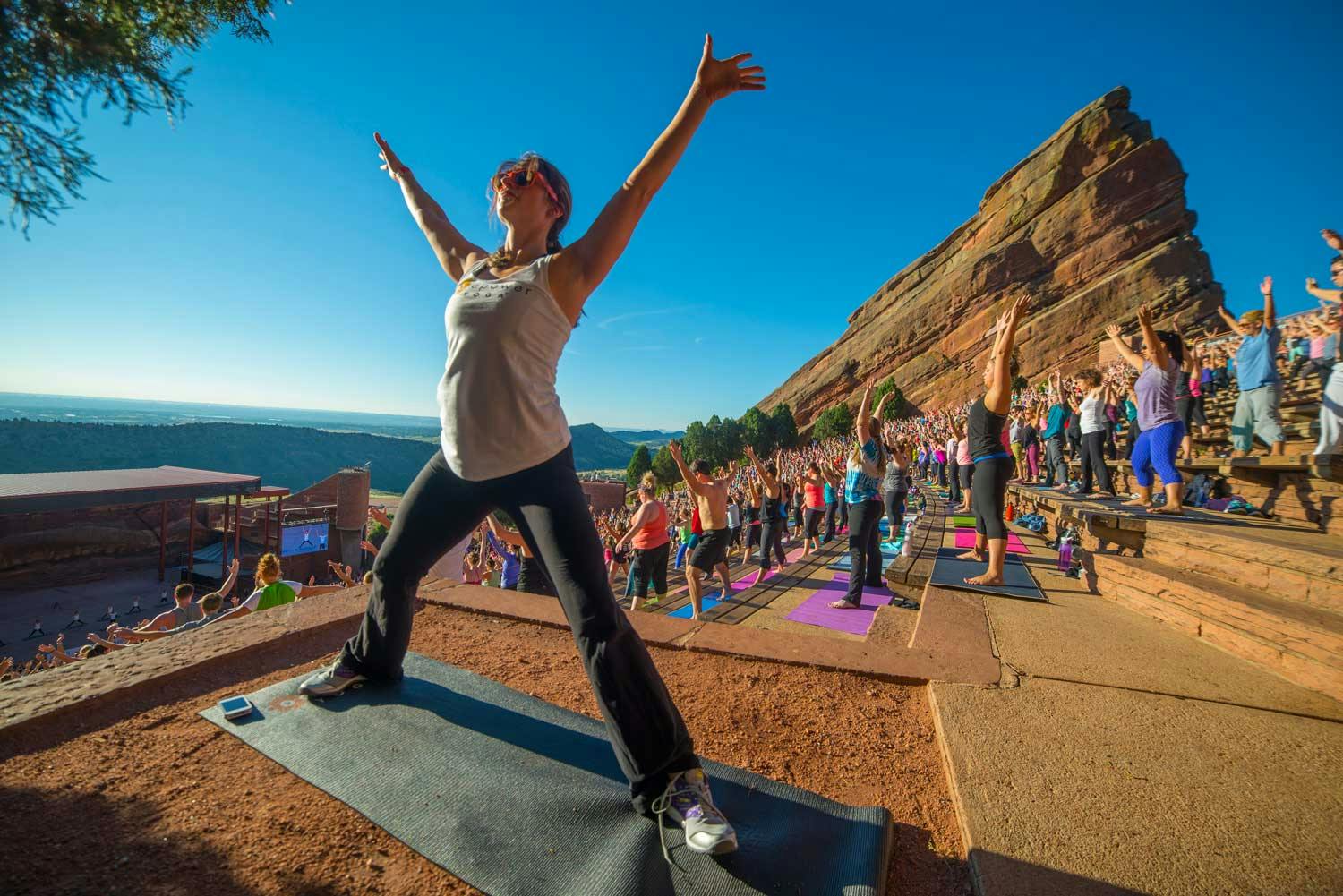 A person strikes a yoga pose reaching for the blue sky at Red Rocks. Behind them on the stairs many people make the same pose, facing the stage at Yoga on the Rocks.