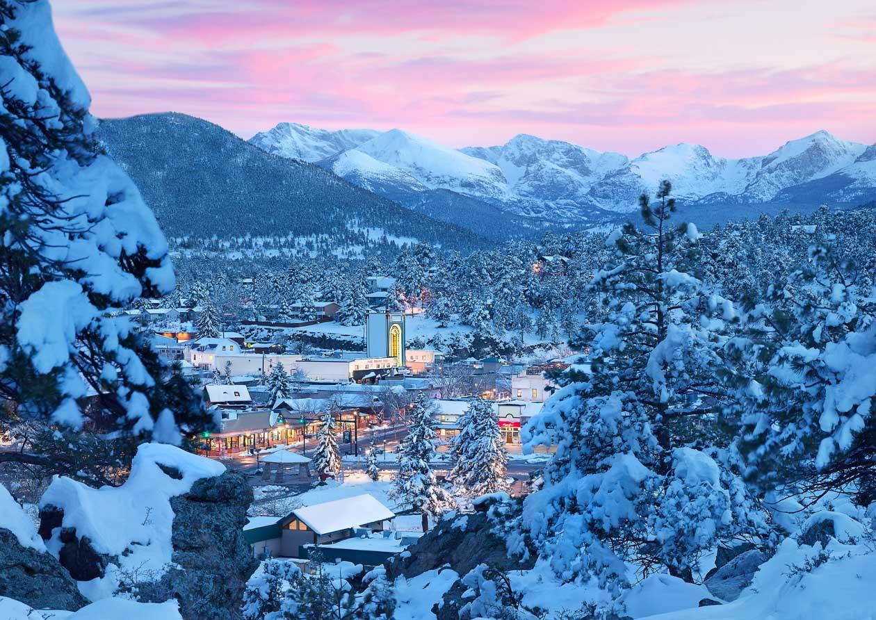 A look down into Estes Park at sunset in the winter. The sky is pink and touches snow-covered mountains and the town is dusted in snow. The surrounding evergreen trees at the viewpoint are coated in thick white snow resembling sugar icing.