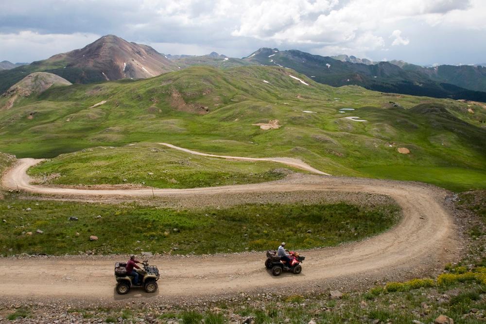 Two red ATVs climb a switchbacking trail along a green mountainside toward a peak's summit