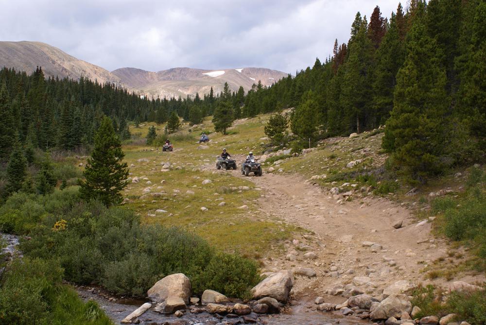 Three ATVs make their way down a rocky dirt path surrounded by evergreen trees and green grass on both sides. In the distance a mountain peak reaches for the blue sky.