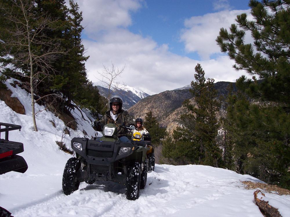 Two ATVs climb a snowy trail with a cliff drop on one side and a snowy hillside on the other