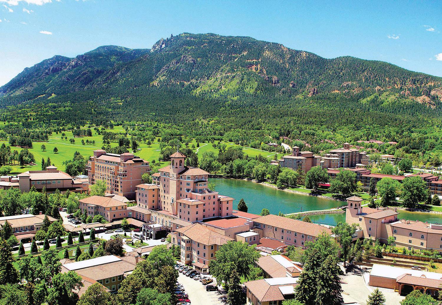 An aerial photo of the Broadmoor in Colorado Springs on a bright, sunny day. The buildings are beige and regal looking, and there is a small body of water sandwiched between the clusters of buildings. In the background are mountainous hills covered in green trees.