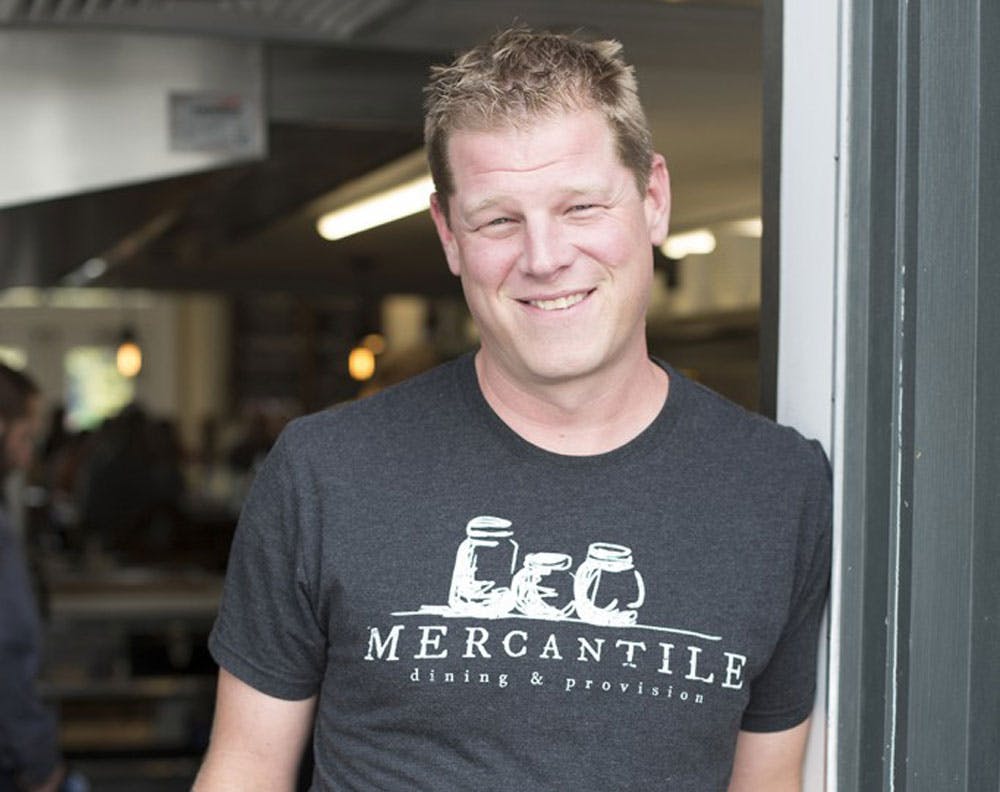 Chef Alex Seidel wears a gray t-shirt and leans against a door jam outside Mercantile Dining & Provisions and Fruition in Denver, Colorado.