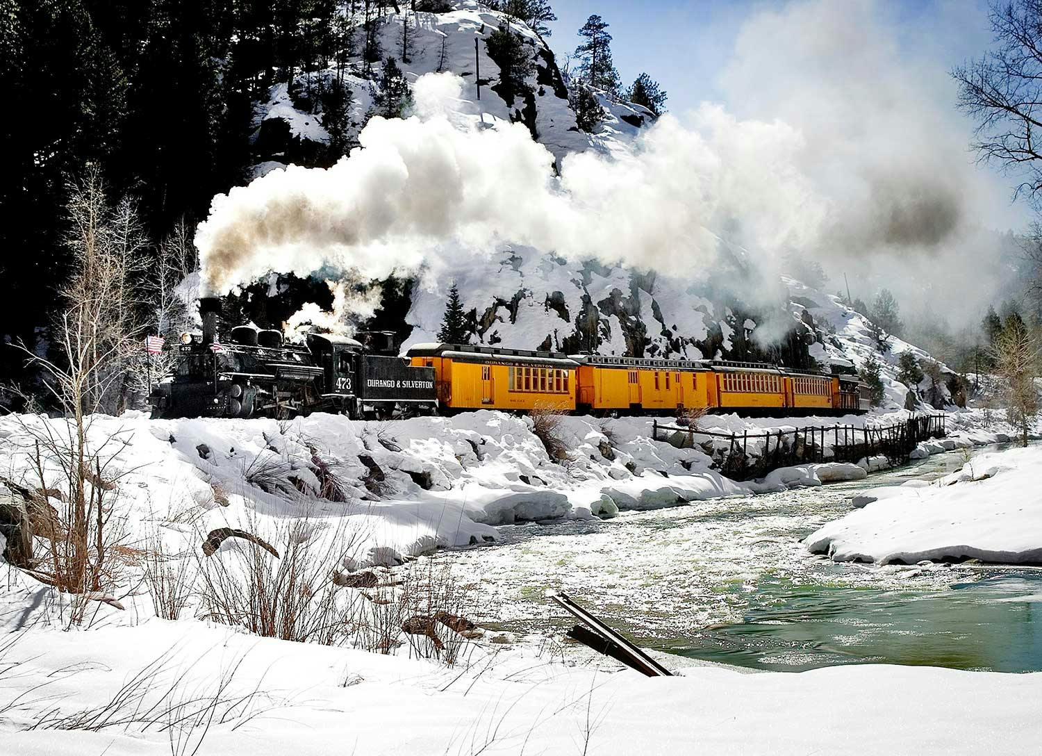 A train moves along a snowy river with mountains all around