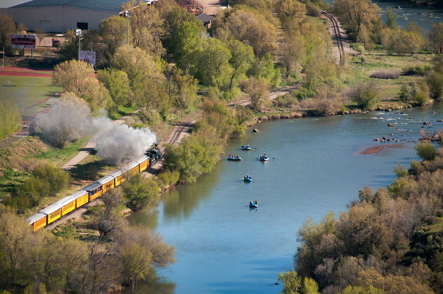 Groups of people in blue inflatable rafts float down a river as a train with yellow passenger cars chugs by on nearby tracks in Colorado.