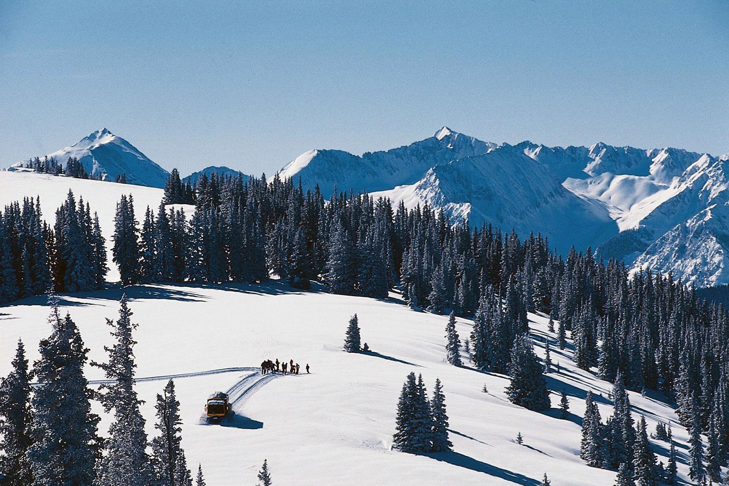 A far away view of snowcat that trundles away from its passengers, who have been left to find fresh tracks down the mountain on a bluebird sunny day