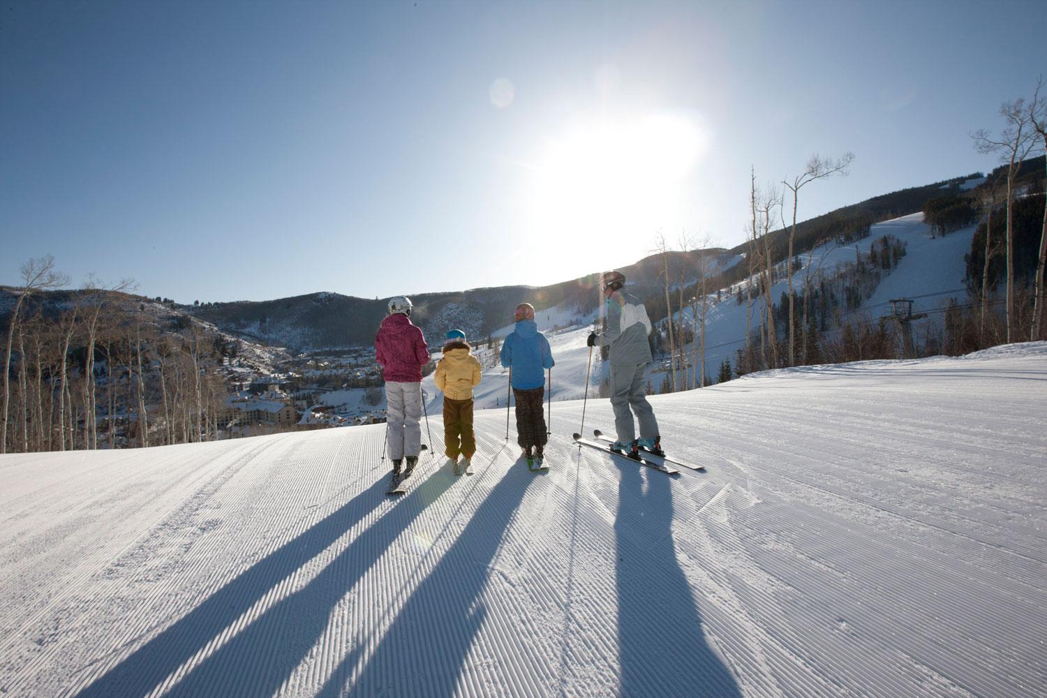 Four skiers are lined up at the top of a slope with the sun shining down on them