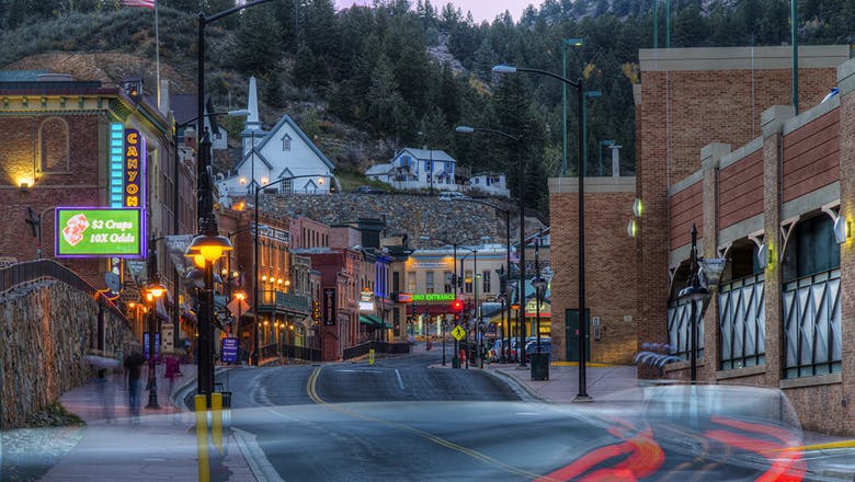 Downtown Black Hawk at sunrise. The street is empty of cars and the neon signs are ablaze. The brick buildings that sit along the winding street are all two to three stories. Up on a hill above the street sits a white church and house. There are evergreen trees above that.