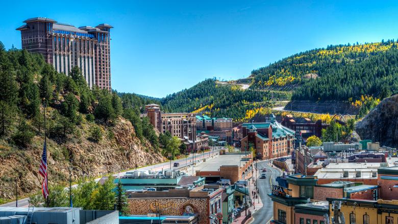 Downtown Black Hawk on a blue-sky fall day. In the distance aspen trees are starting to turn golden, and a winding road is making its way up a mountain. The winding Main Street has a couple cars driving by historic brick buildings. On the left a hulking building is being built above town. 