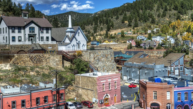 Historic, downtown Black Hawk on a bright, blue-sky summer’s day. Evergreen trees are in the background, covering mountains. Along a stone wall, a light blue historic church sits above old Western two story buildings. 