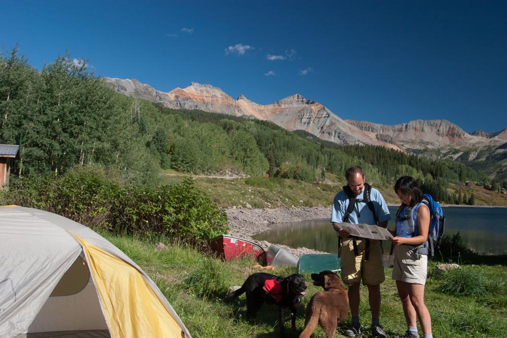 A couple wearing backpacks cosult a map near a red canoe, a tent and a black lab.