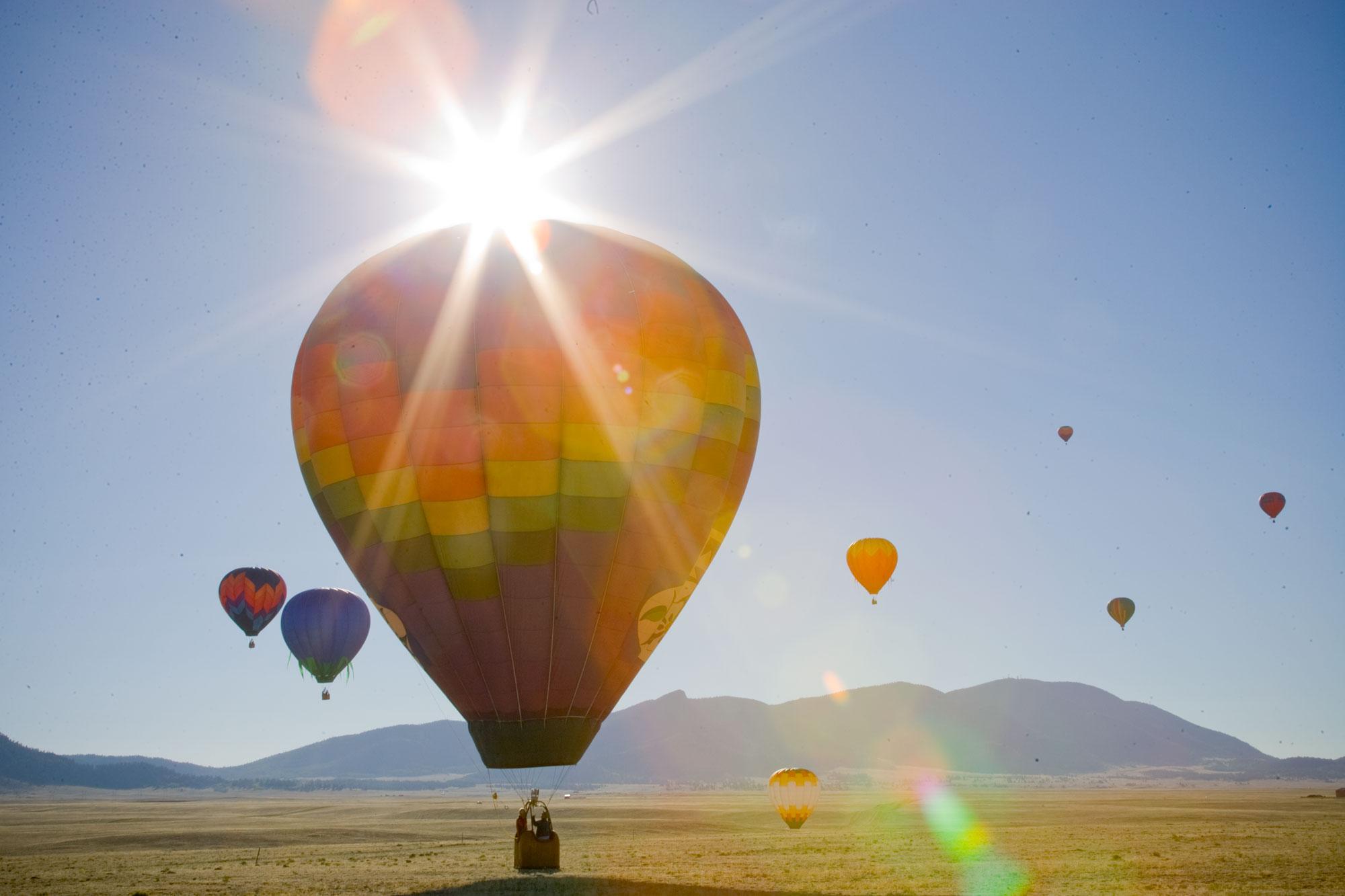 We see a rainbow-colored hot-air ballon with a burst of sunlight over it; in the distance, six or seven other balloons hover in the air