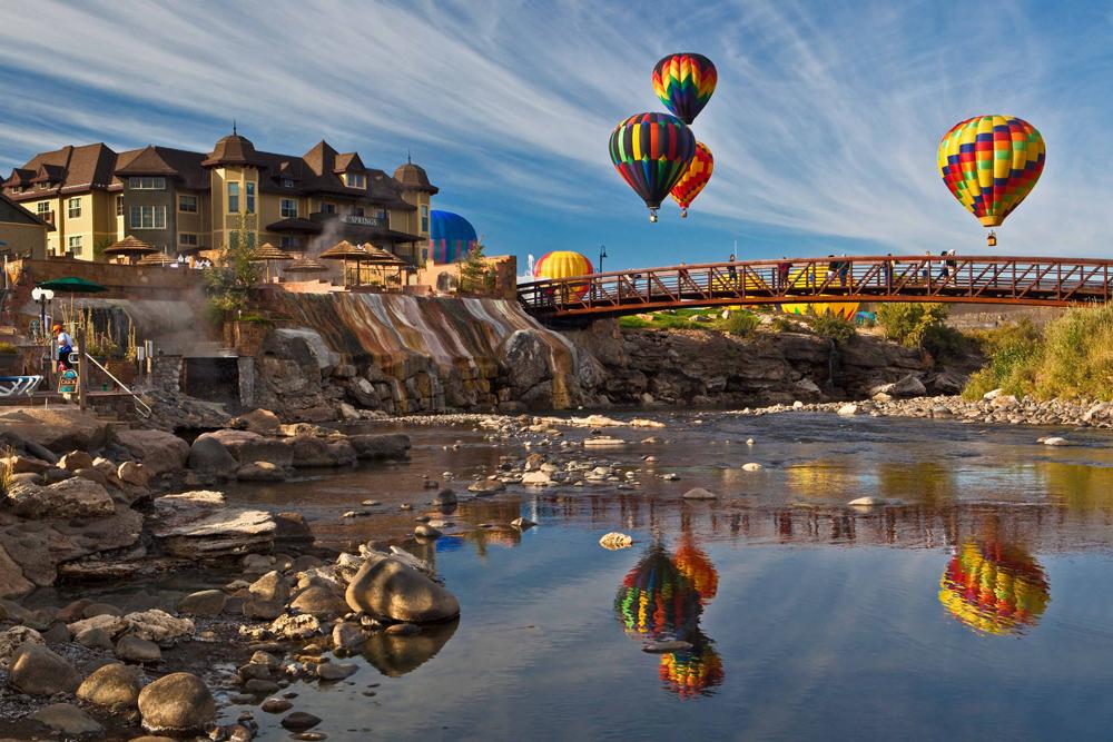 Four rainbow-colored hot-air balloons hover over a bridge, hotel and river. Their shapes and colors are reflected in the water