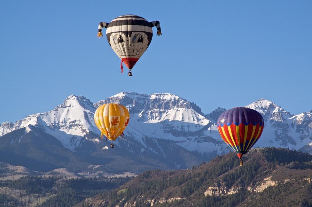 Three hot air balloons decorated soar above an evergreen covered valley with snowcapped mountains in the distance. One balloon is blue, red, yellow and green stripes. One is yellow with fall leaves on it. And the other is black and white with a face on it.