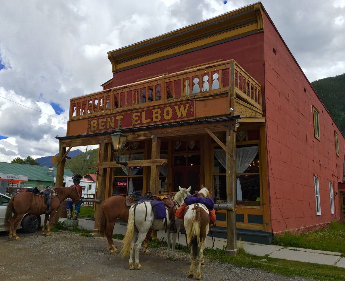 Four horses are tied up outside of a historical Western building the has a sign that says "Bent Elbow" across the balcony.