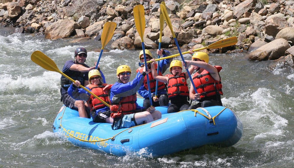 A group of people wearing helmets and life jackets hold up yellow paddles and cheer as the float along the whitewater rapids near Idaho Springs, Colorado.