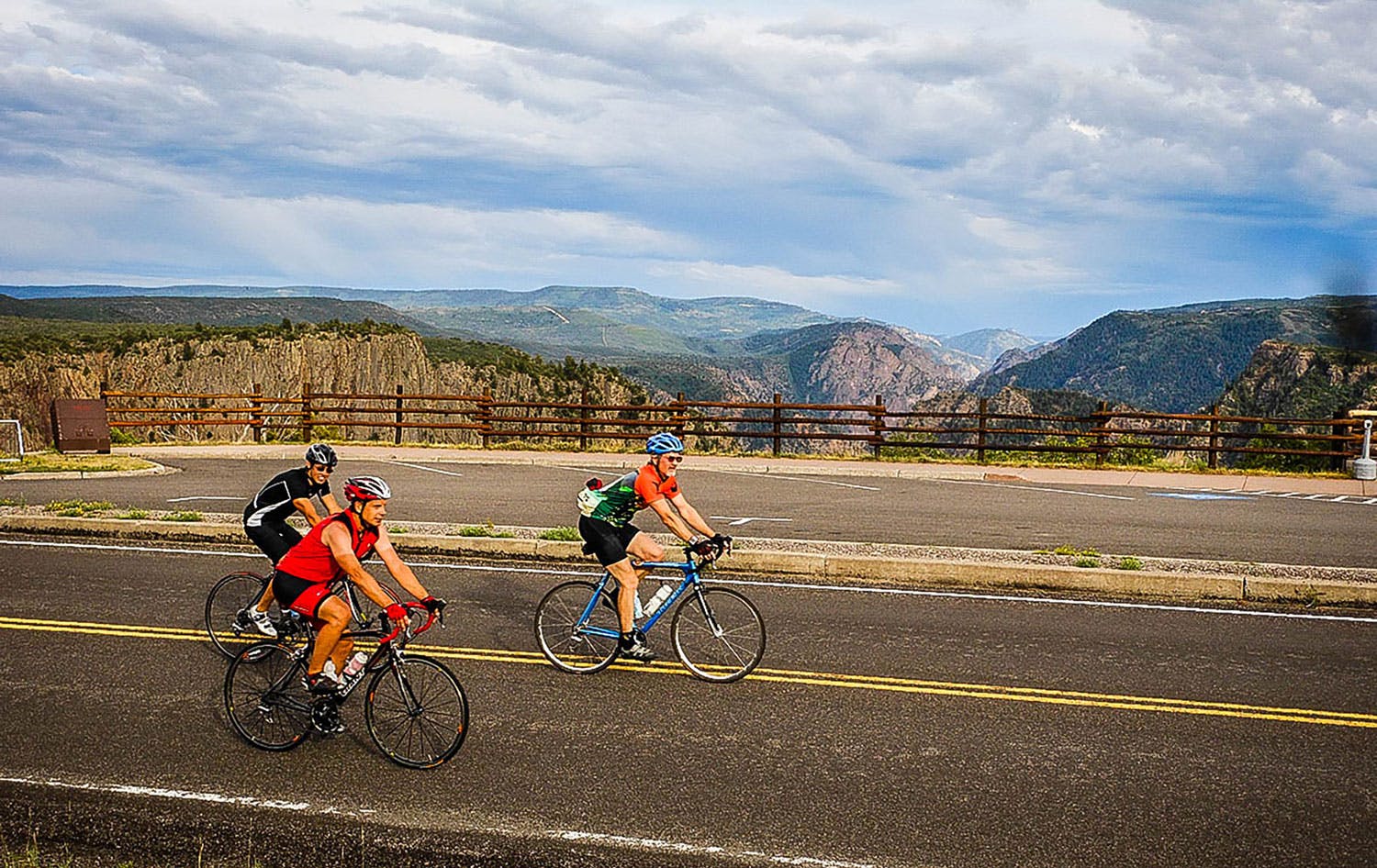 Three road cyclists ride over a blacktop roads with Black Canyon of the Gunnison National Park as their backdrop.