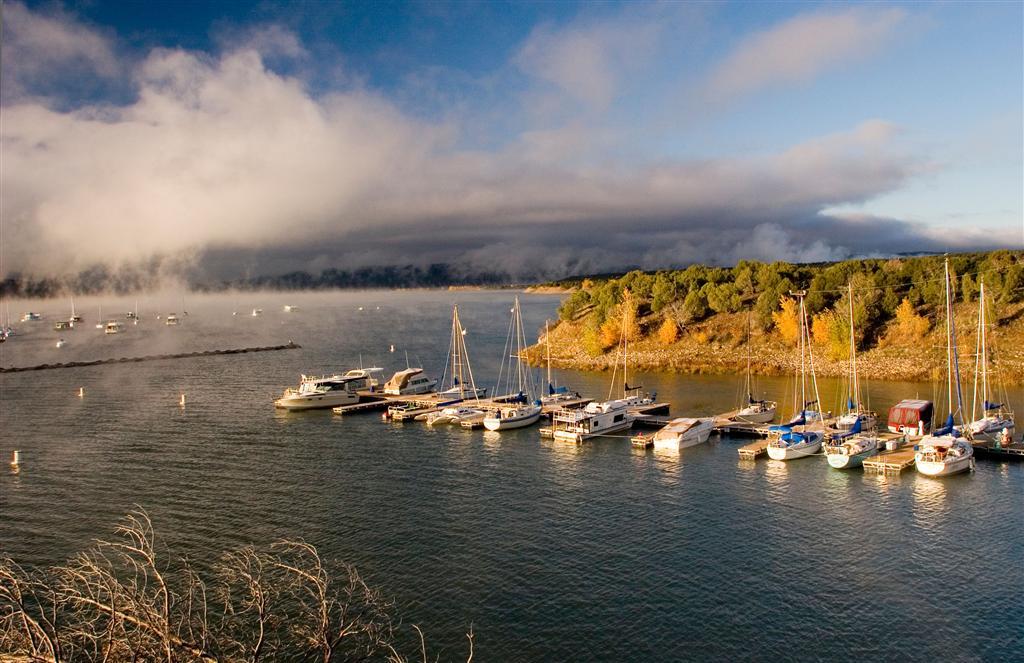 A row of sailboats is docked along a pier on a lake with mountain peaks in the background