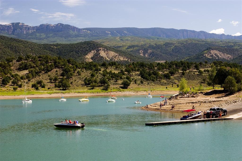 Multiple boats move through the teal water at Ridgway State Park with green trees on hills in the background.