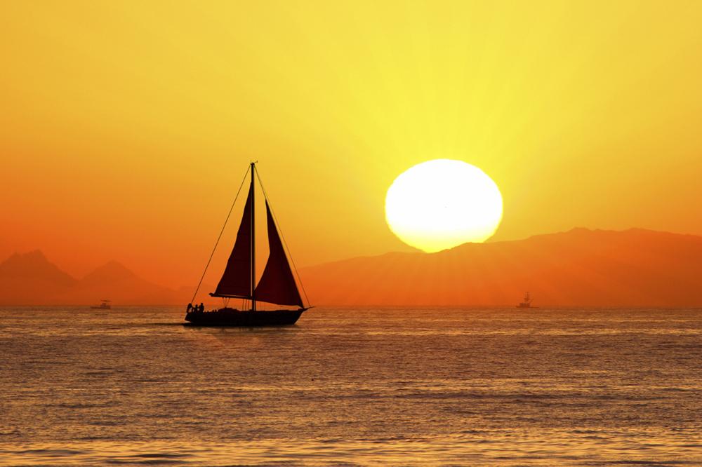 A sail boat coasts across the water at sunset. The sky is yellow with the sun a big circle going behind orange-hued mountains.