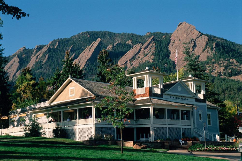 A woodframe building with wrap-around balcony sits in a green meadow between Boulder's famous Flatirons, wedge-shaped mountains