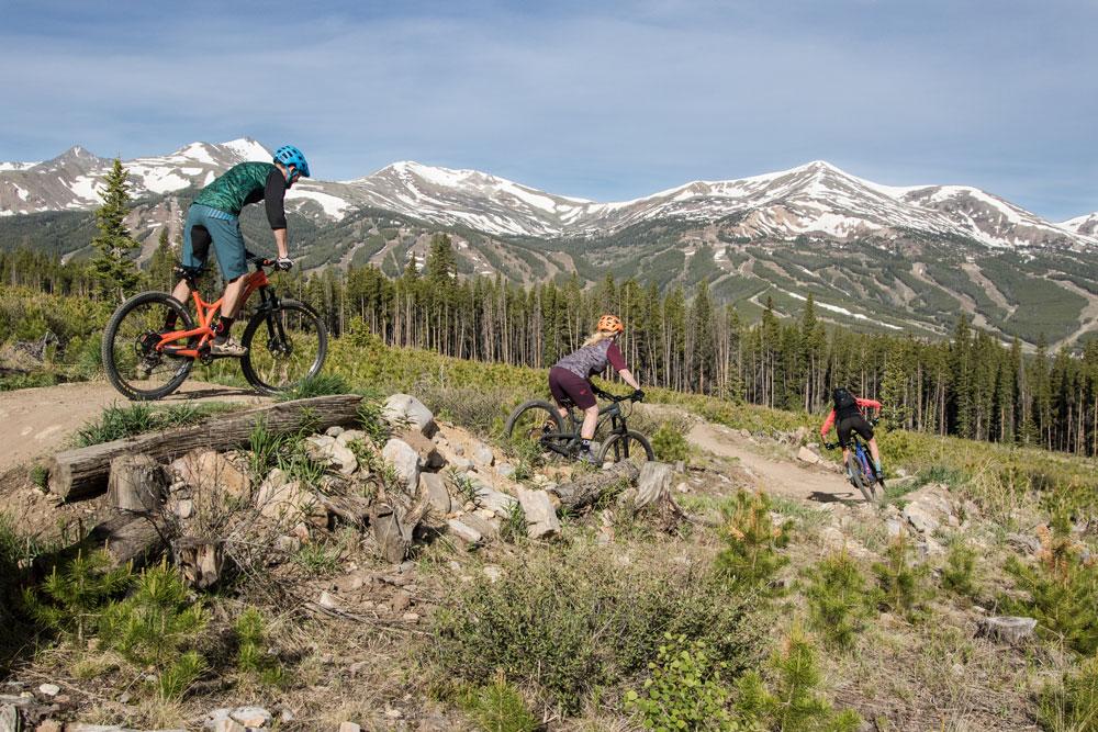 Three people mountain bike on a summer day down a single track. The sky is a hazy blue and there are snow-capped mountains in the background.