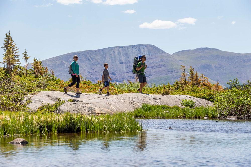 A family with a child hikes near a blue lake during summer. There is a light blue sky with a few whips clouds and mountains are in the background.