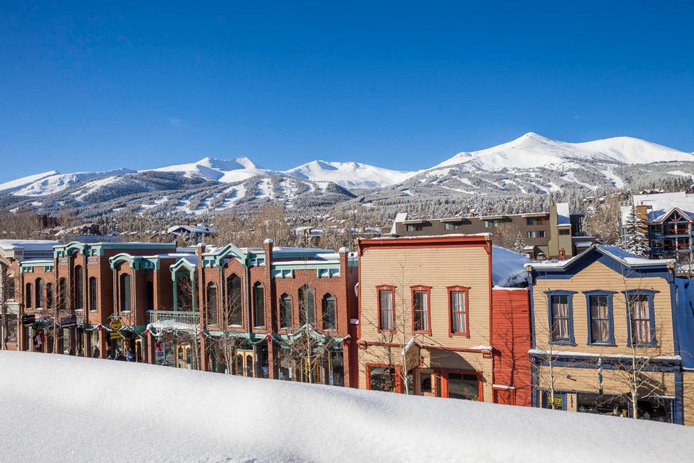 Historic Western Breckenridge sits beneath a bright-blue sky with snow-covered mountains in the background.