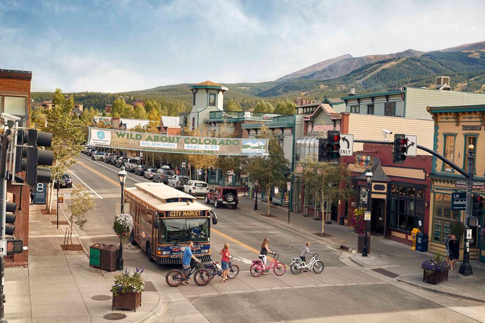 Four people walk their bikes across the street in front of a public bus in downtown Breckenridge. The Western storefronts sit below mountain ski runs. 