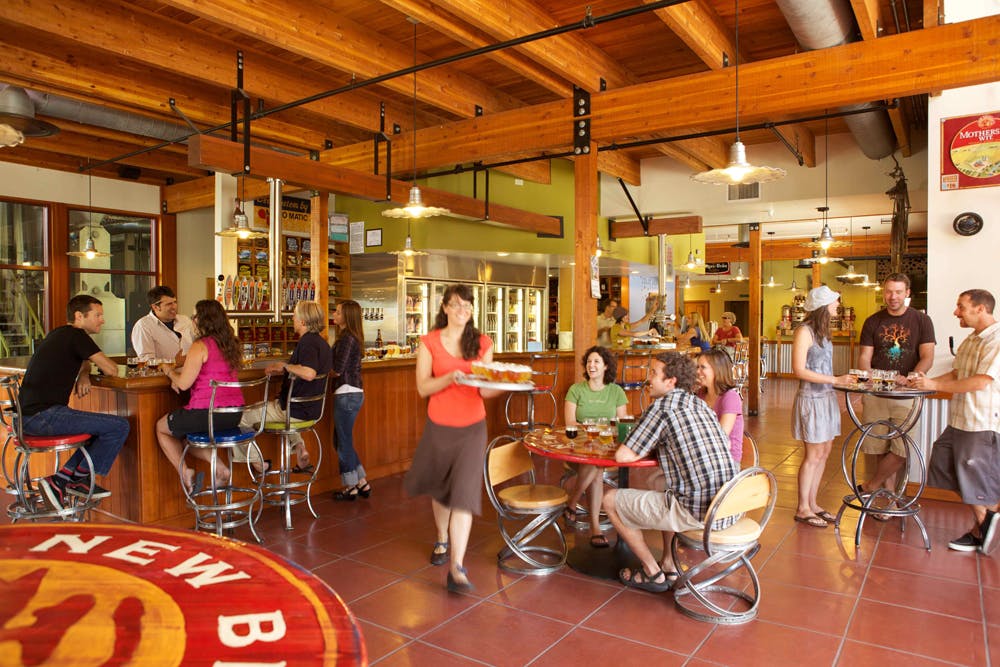 A waitress carries a tray of beers through the tasting room, which has the Fat Tire logo on the floor. Several tasters sit at tables