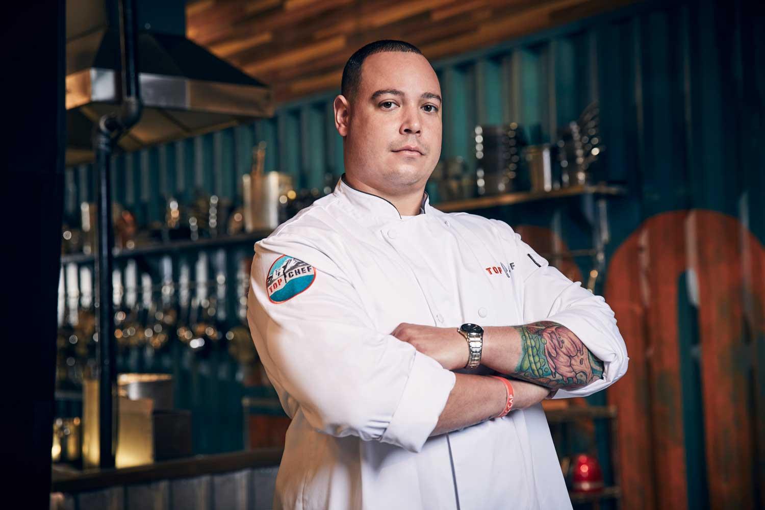 Top Chef Colorado Cheftestant Brother Luck stands in his restaurant in his chef's coat with arms crossed