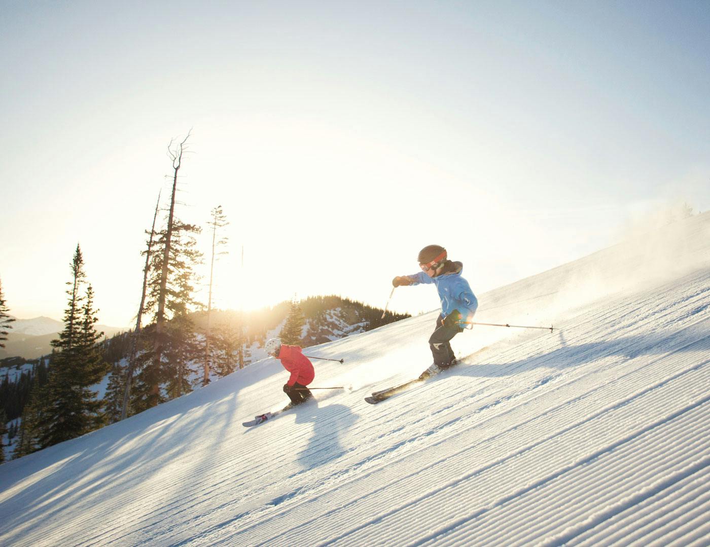 An adult and child swish down a snow run lined with corduroy as the sun sets behind them