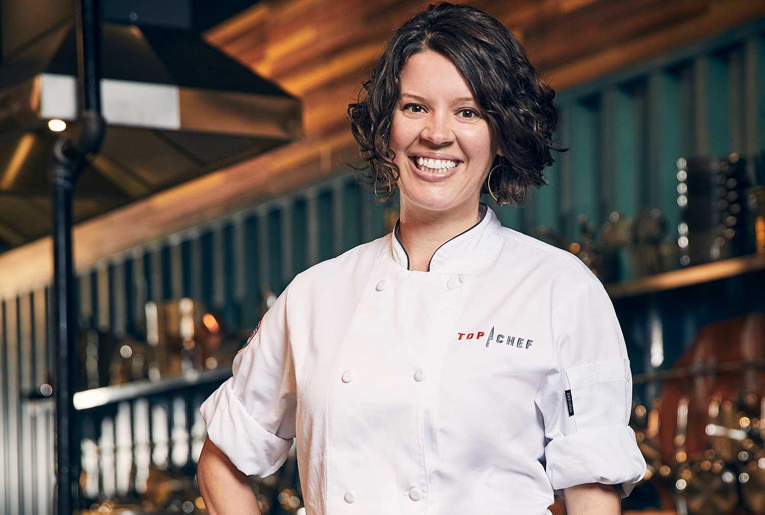 Top Chef Colorado Cheftestant Carrie Baird smiles big and poses in her white Top Chef coat.