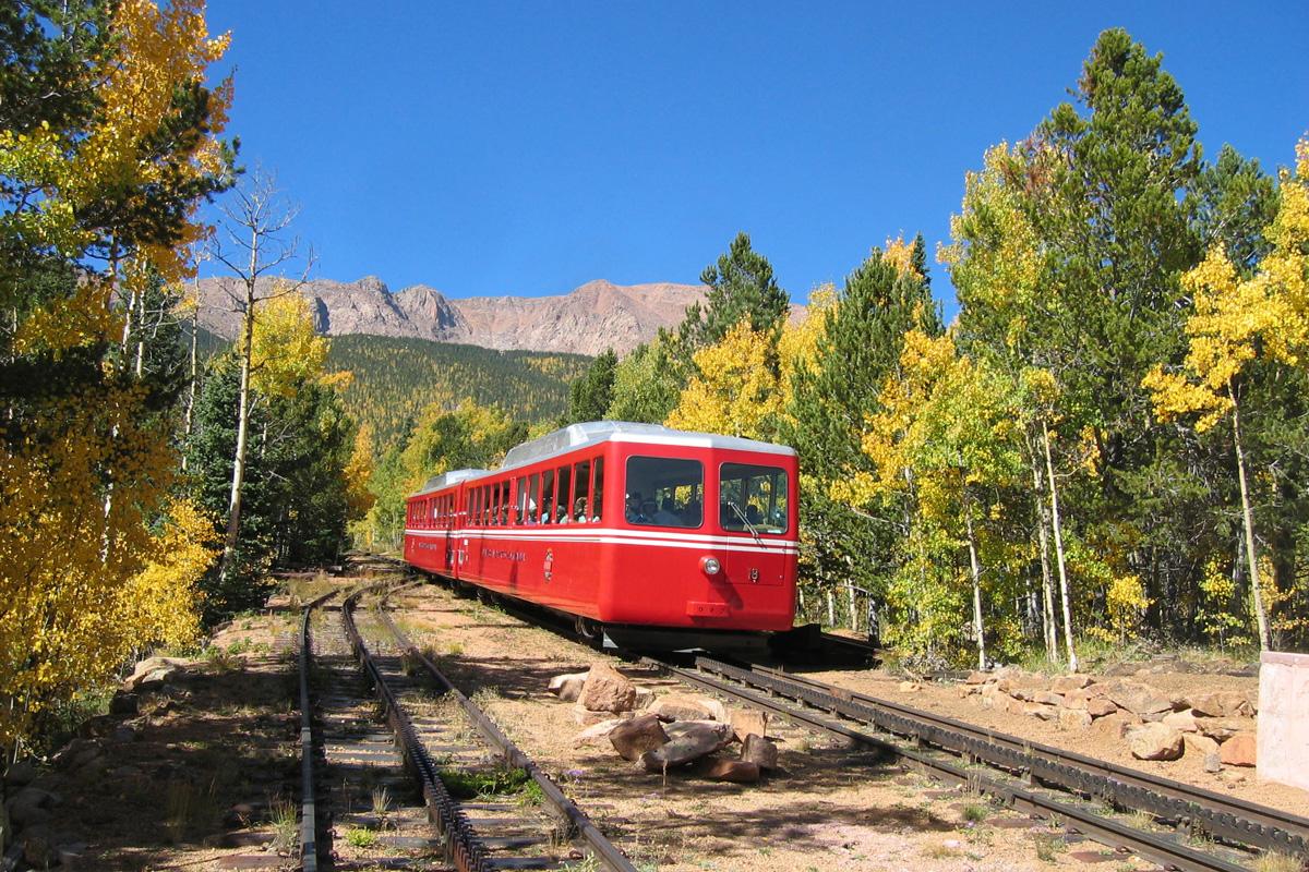 A red train with a double white-stripe design and a boxy appearance drives down a set of railroad tracks in Colorado. Aspens and evergreens line both sides of the track and mountain peaks rise up in the distance.