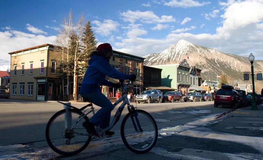 A cyclist rides through a crosswalk near historic storefronts in Crested Butte, Colorado. In the background, a snow-capped mountain rises about the town buildings.