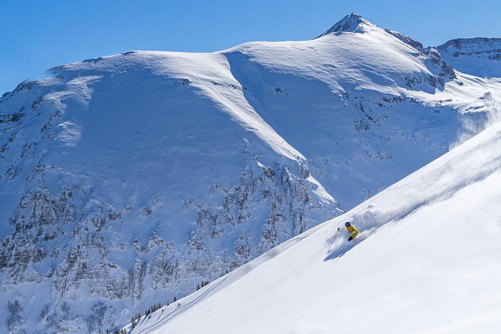 A person skis down a strep slope with snow-covered mountains in the background.