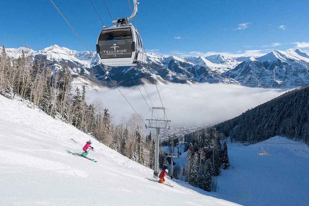 Two skiers ski under a Telluride-labeled gondola. There are snow-covered peaks in the background.