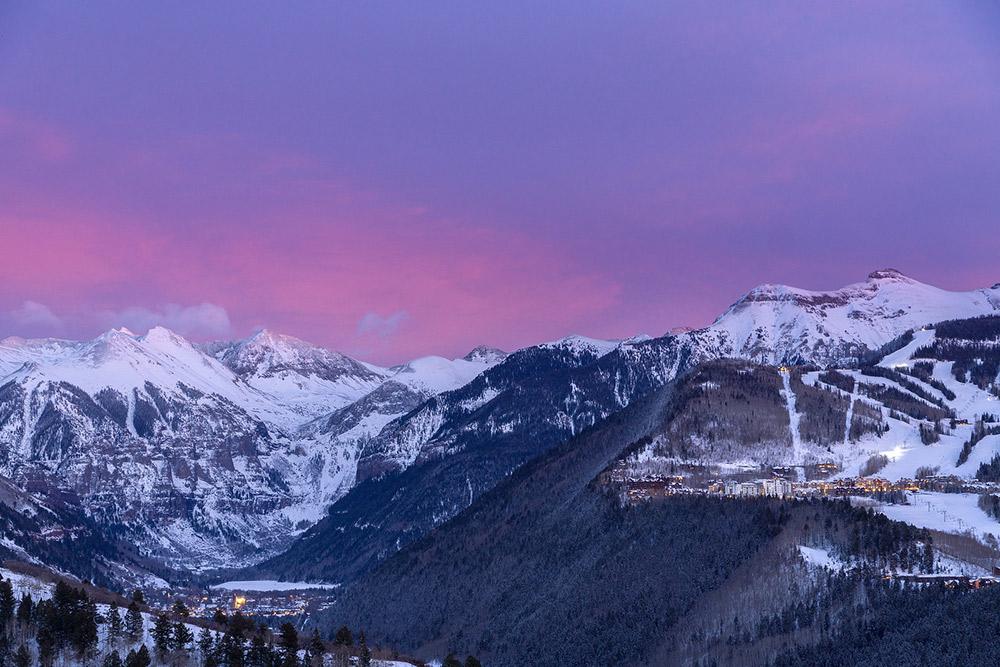 A pink and purple sky sit above a snow-covered mountain range with a town in the valley below it.