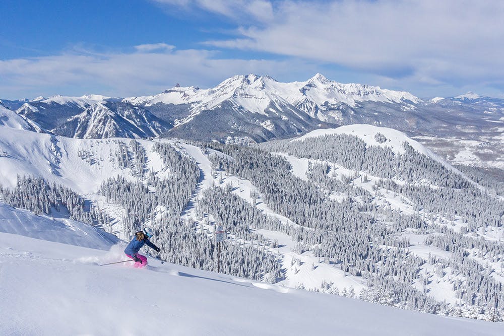 A skier skis down a snowy mountain pass with snowy paths and evergreen trees dusted in snow. There's mountain peaks covered in snow in the distance.