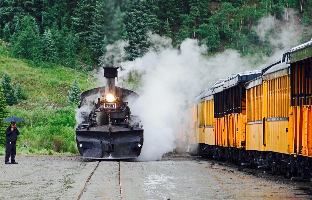 A black, steaming engine car passes yellow train cars of the Durango & Silverton Narrow Gauge Railroad