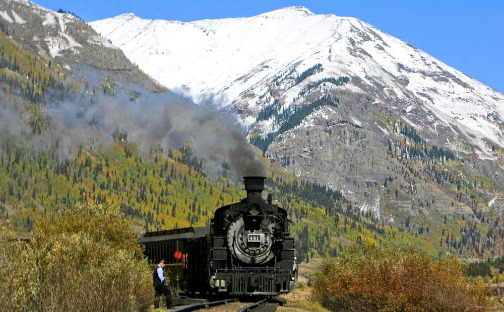 A black train engine chugs its cars up the tracks with a snow-covered peak in the background