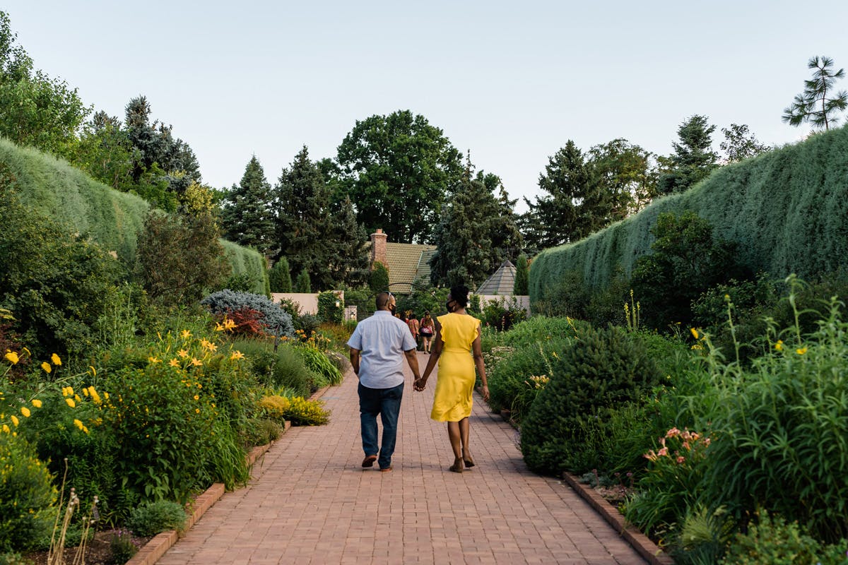 A couple holds hands and walks down the middle of an orange-paver path under a light blue sky at Denver Botanic Gardens. On both sides are green vegetation with yellow flowers.