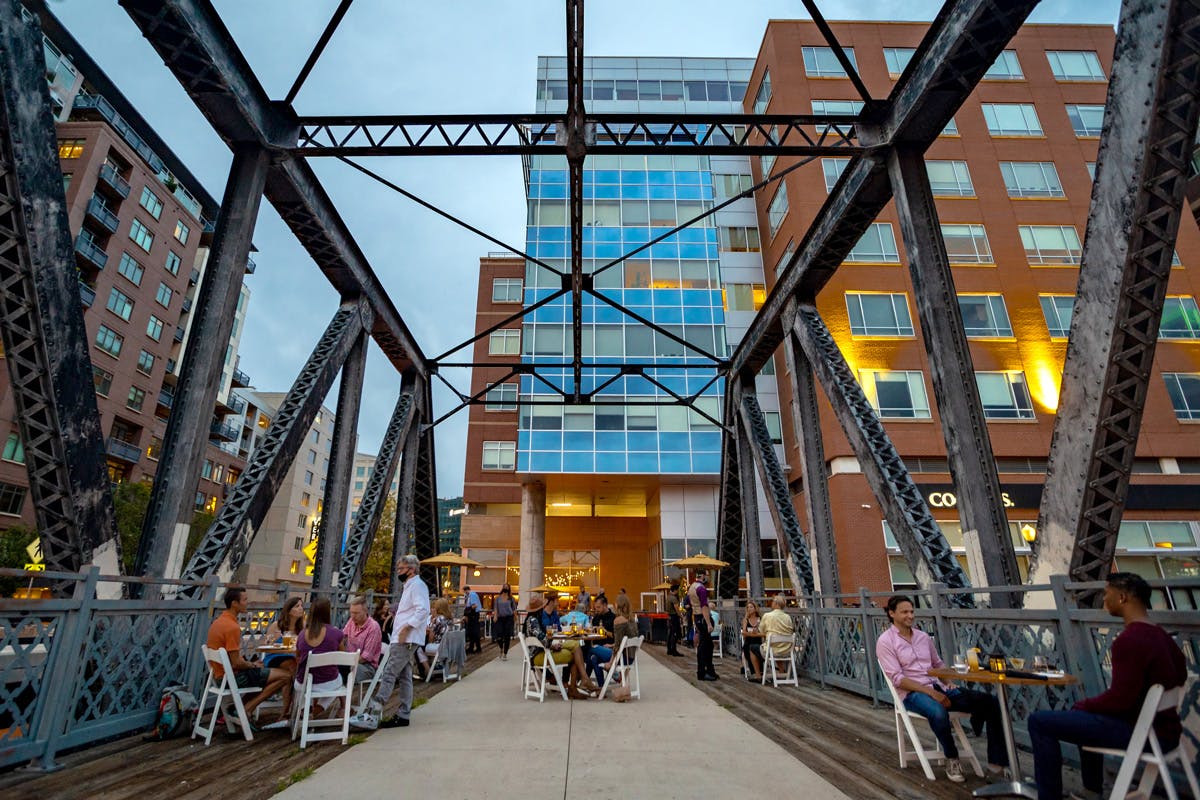 People sit outside at white tables on an old railroad bridge at dusk. In the background a glass and brick multi-store building hovers over.