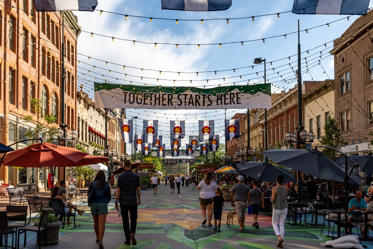 People walk down Larimer Square with a sign that says "Together Starts Here." Twinkle lights are strung up between red-brick buildings and banners with the Colorado flag in the distance.