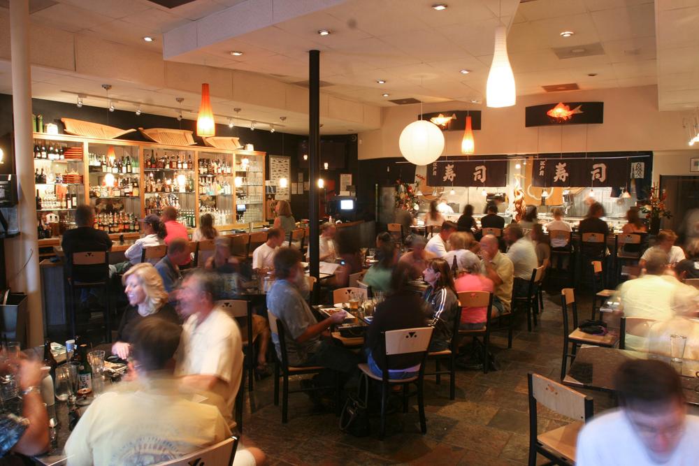 The crowded dining room of a restaurant; the bar is stocked with bottles and you can see chefs working in the kitchen beneath banners with Japanese characters on them