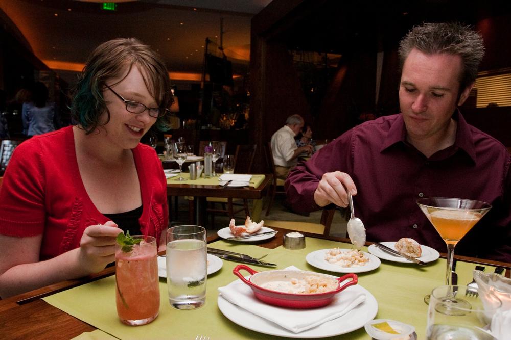 A couple sits at a fine-dining restaurants, both digging into their meals. A martini and cocktail glass sits next to their plates