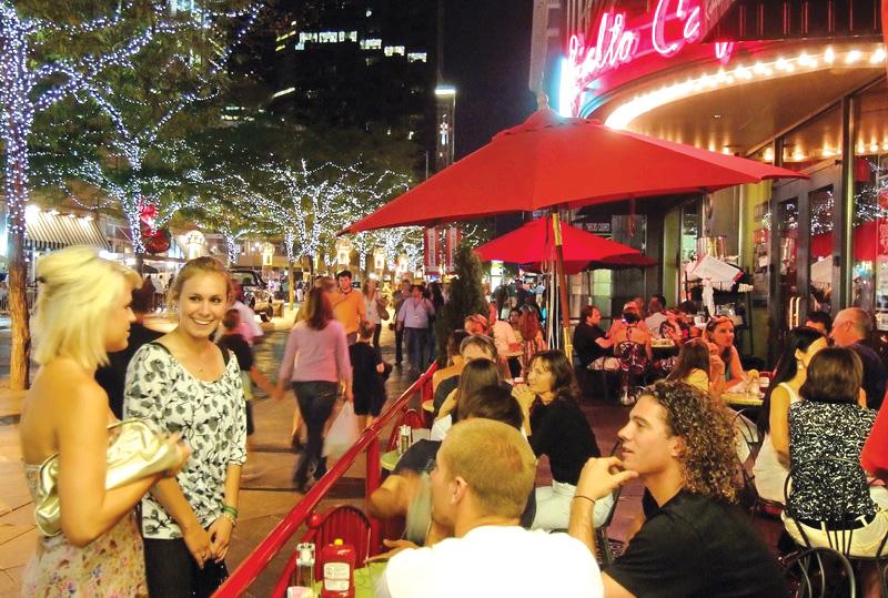 Diners and pedestrians chat under the awning of the Rialto Cafe; white lights are draped in the trees behind them