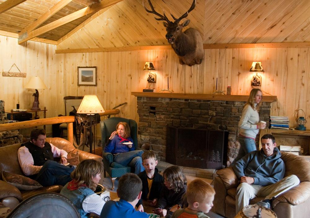 Inside a lodge, guests are gathered in front of a fireplace in chairs and couches reading and playing games. The walls are covered in light-colored wood and the head of a trophy elk is mounted above the fireplace